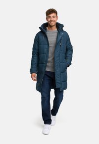 Tyrkis pufferjakke med hette og glidelås, med en quiltet design. Brukt over en grå genser og mørke jeans med hvite sneakers.