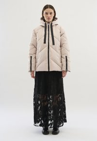 Lichtroze fluffy jas met capuchon, geribbeld textuur en chevronpatroon; gecombineerd met een zwart kanten maxi-jurk. Zwarte accenten en ritsen.