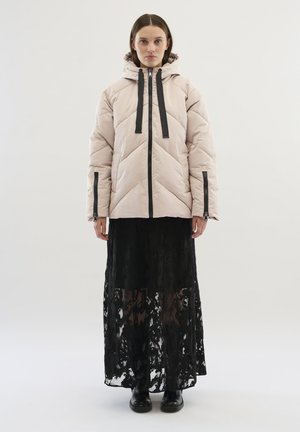 Lichtroze fluffy jas met capuchon, geribbeld textuur en chevronpatroon; gecombineerd met een zwart kanten maxi-jurk. Zwarte accenten en ritsen.