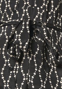Vestido de tela negra con patrón de lunares blancos, que cuenta con una cintura atada, detalles de botones y una textura fluida. La vista en primer plano enfatiza el diseño.