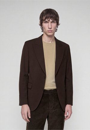 Ung mand med bølget brunt hår iført en mørkebrun blazer, beige skjorte og mørkebrune kordfløjspants, stående mod en ensfarvet baggrund.