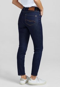 Jeans ajustados de mezclilla azul oscuro. Presentan una cintura alta, costuras naranjas y un parche de cuero de la marca. Combinados con zapatillas blancas.
