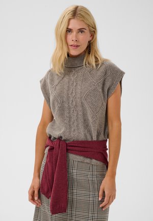 Femme blonde portant un pull gris sans manches à col roulé, un pull bordeaux noué autour de la taille et une jupe à carreaux.