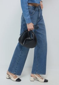Jeans en denim bleu à jambes larges, sac en cuir noir au design arrondi, et escarpins beiges avec bout noir et talon bloc.