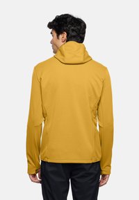 Giacca gialla con cappuccio realizzata in tessuto liscio, caratterizzata da una vestibilità comoda e cuciture posteriori per la forma. Nessun hardware o logo visibile.