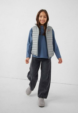 Fille souriante marchant, portant un gilet matelassé gris clair sur un chemisier bleu à manches longues, un jean noir à jambes larges et des chaussons gris.