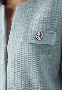Gros plan d'une veste en tissu bleu clair texturé avec des motifs verticaux, comportant une poche à rabat et un bouton en argent brillant de forme irrégulière.