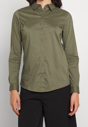 Femme portant une chemise boutonnée vert olive à manches longues et un pantalon noir, debout devant un fond clair uni.