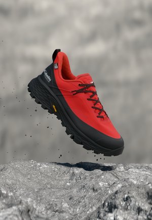 Κόκκινο και μαύρο ορειβατικό παπούτσι με ανθεκτική σόλα και μαύρα κορδόνια, στον αέρα πάνω από βραχώδες έδαφος, προωθώντας την αντοχή και την εξωτερική χρήση.
