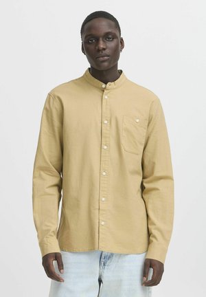 Camisa de botones de manga larga, color marrón claro, con un cuello redondeado y un bolsillo en el pecho. La tela parece tener textura y ser suave.