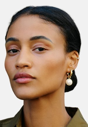 Goldene Hohrringe mit einer Perlenakzent, glatte Oberfläche und runder Form. Dezentes, natürliches Make-up und ein neutrales Shirt sind sichtbar.