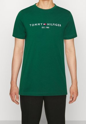 Zielona koszula z krótkim rękawem z logo "Tommy Hilfiger EST. 1985" i emblematem flagi na piersi, noszona przez stojącą osobę.