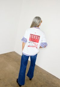 Άτομο με μακριά γκρίζα μαλλιά φοράει λευκό T-shirt με γραφικό rodeo πάνω από ριγέ πουκάμισο και μπλε τζιν καμπάνα, στέκεται σε τσιμεντένιο πάτωμα δίπλα σε λευκό τοίχο.