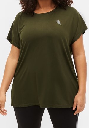 Kvinde iført løs, olivengrøn kortærmet skjorte med lille hvidt logo på brystet og sorte leggings.