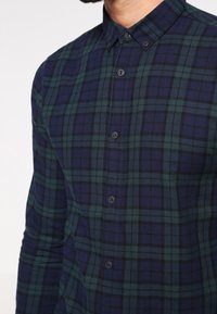 Geruite flanellen blouse in donkerblauwe en groene tinten, met een button-down kraag en een gestructureerde stof met ruitpatroon.