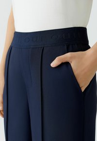 Navy pleated pants with a textured waistband featuring an embossed "OUI" logo, side pocket, and tailored design for a fitted look.