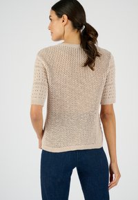 Un haut en tricot beige à manches courtes avec un motif de treillis ouvert et un ourlet côtelé. Porté avec un jean fitted bleu foncé. Vue arrière présentée.