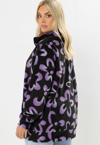 Kurtka polarowa z czarnym tłem i fioletowymi kwiatowymi wzorami. Wysoki kołnierz, długie rękawy i luźny krój.