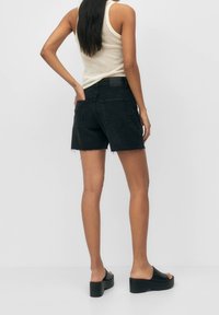 Mujer con cabello largo que lleva una blusa sin mangas blanca, pantalones cortos de mezclilla negra con bordes deshilachados y sandalias de plataforma negras, apoyada contra una pared lisa.
