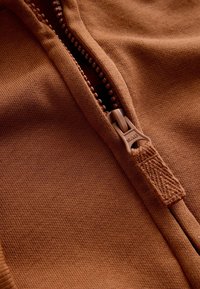 Brown zipped fabric featuring a textured surface with a smooth zipper and a matching pull. Close-up view emphasizes stitching and material.