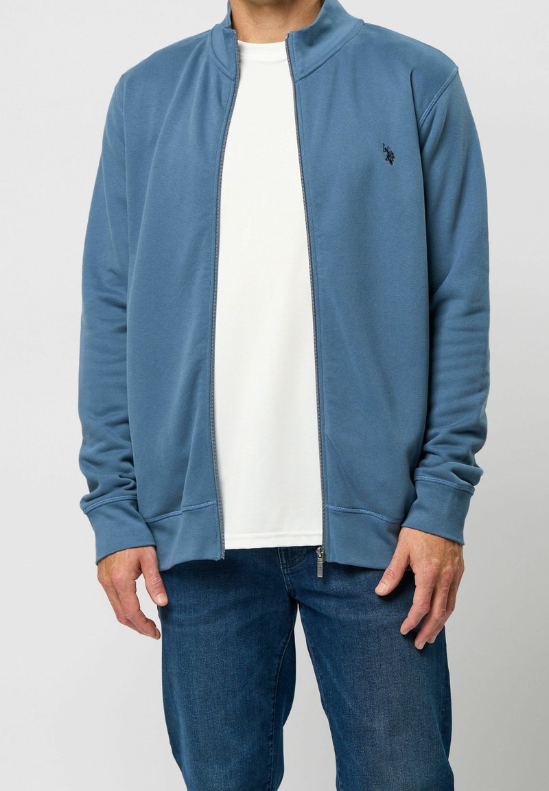 Homme portant une veste bleue zippée par-dessus une chemise blanche et un jean bleu, debout avec les mains détendues le long du corps.