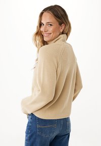 Beige pletený svetr s vysokým límcem a žebrovanou strukturou, nošený přes modré denimové džíny. Design má spadlé ramena a boční rozparky.