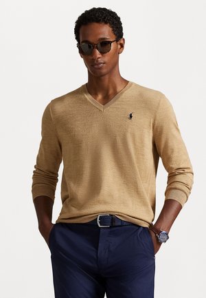 Pull beige à col en v avec un tricot texturé, doté d'un petit logo bleu marine sur la poitrine. Assorti à un pantalon bleu marine foncé et une ceinture marron.