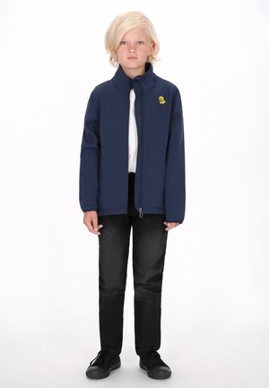 Giacca softshell blu navy con colletto alto e logo giallo, indossata sopra una camicia bianca, abbinata a pantaloni neri e sneakers nere.