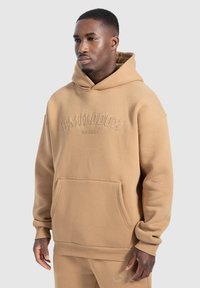 Sudadera marrón hecha de tela suave, con un bolsillo tipo canguro, puños acanalados y un logo elevado en el pecho. Diseño simple y casual.