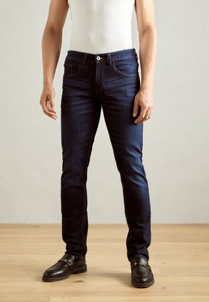 Man die donkere, donkerblauwe slim-fit jeans en zwarte leren loafers draagt, staand op een houten vloer tegen een effen lichte muur.