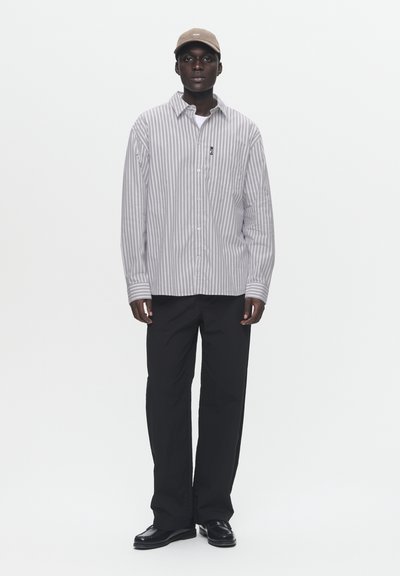 Gestreiftes Hemd in Grau und Weiß, kombiniert mit schwarzen Hosen und dunklen Schuhen. Eine beigefarbene Mütze ergänzt das Outfit.