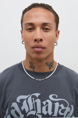 Model iført en grå t-shirt med store hvide bogstaver, en perlehalskæde med sølvdetaljer og synlige tatoveringer af en rose og en sommerfugl.