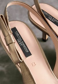 Gold metallic slingback heels with a smooth, pale pink insole. Features a bow accent and a narrow heel. Branding visible on the insole.