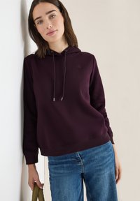 Frau mit schulterlangem braunem Haar, die einen dunkelvioletten Hoodie und blaue Jeans trägt. Sie lehnt gegen eine weiße Wand und hält einen olivgrünen Gegenstand.