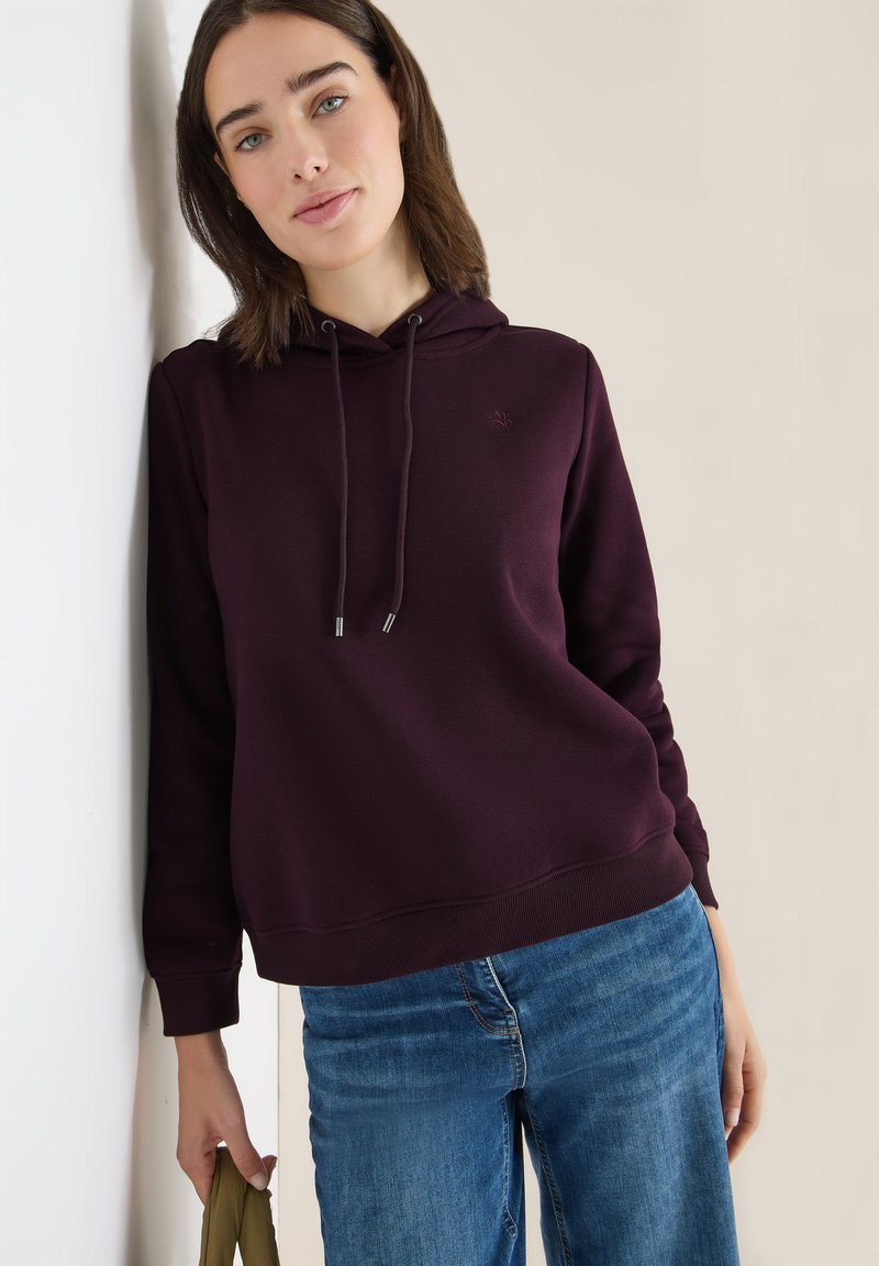 Frau mit schulterlangem braunem Haar, die einen dunkelvioletten Hoodie und blaue Jeans trägt. Sie lehnt gegen eine weiße Wand und hält einen olivgrünen Gegenstand.