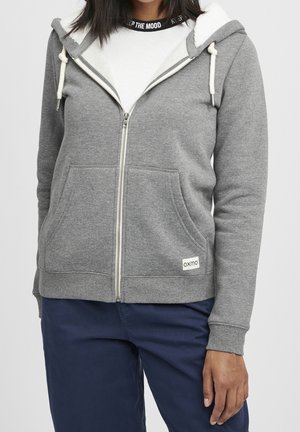 Personne portant un sweat à capuche gris zippé avec une doublure en polaire blanche, une chemise blanche et un pantalon bleu marine, visible des épaules aux hanches.