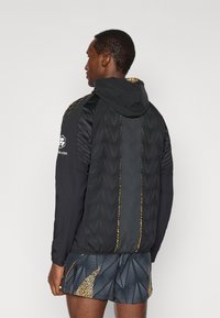 Μαύρο αθλητικό μπουφάν με κουκούλα, με υφασμάτινες επιφάνειες και χρυσές λεπτομέρειες. Συνδυασμένο με σορτς με μοτίβα σε σκούρους τόνους.