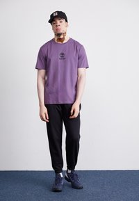 Violetinis medvilninis marškinėliai su Timberland logotipu, juodi treniruotėms skirti kelnių modeliai ir tamsūs batai. Modelis stovi ant mėlyno kilimo prieš paprastą foną.