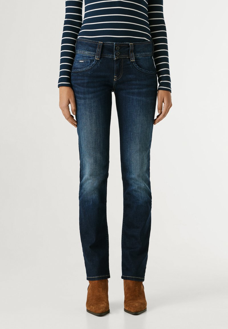 Persona che indossa jeans in denim blu scuro, una maglietta a maniche lunghe a righe blu navy e bianco e stivaletti in camoscio marrone, in piedi dritta contro uno sfondo semplice.