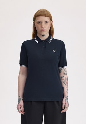 Femme aux cheveux rouges et tatouages portant un polo noir avec des rayures blanches sur le col et les manches, posant devant un fond uni.