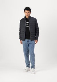 Veste matelassée grise avec un col montant, associée à un t-shirt noir et un jean clair. Des sneakers blanches complètent l'ensemble.