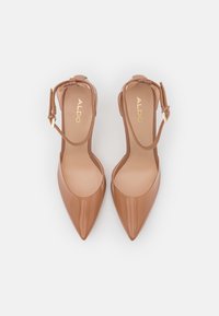 Kožené lodičky v béžové barvě s vysokým podpatkem a špičatou špičkou, doplněné o řemínek kolem kotníku se zlatým sponou. Hladký povrch, minimalistický design.