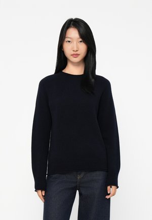 Ung kvinne med rett svart hår iført en mørk marineblå genser med lange ermer og mørke jeans, stående mot en hvit bakgrunn.