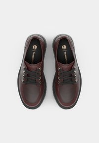 Pantofi din piele de culoare burgundă, cu șireturi negre, gauri metalice și o talpă texturată de culoare neagră. Branding vizibil pe insole.