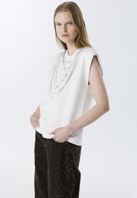 Top sin mangas blanco con diseños de cadenas metálicas en capas, combinado con pantalones de estampado de leopardo marrón. Tejido suave con un ligero brillo.