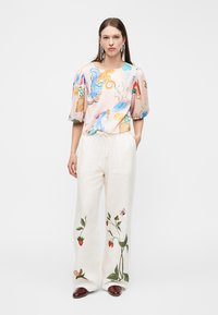 Femme en blouse légère ornée de créatures marines abstraites colorées et pantalon blanc avec des fraisiers brodés, se tenant devant un fond blanc.