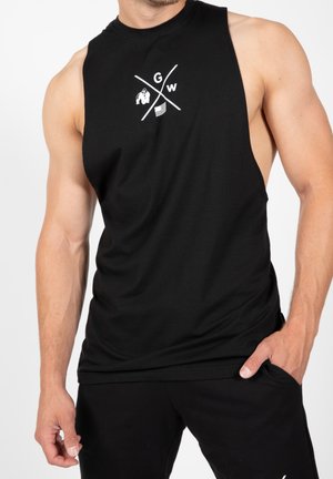 Muskularny mężczyzna ubrany w czarny bezrękawnik z emblematem złożonym z krzyżujących się linii, przedstawiającym goryla, literę G, W oraz symbole flag.