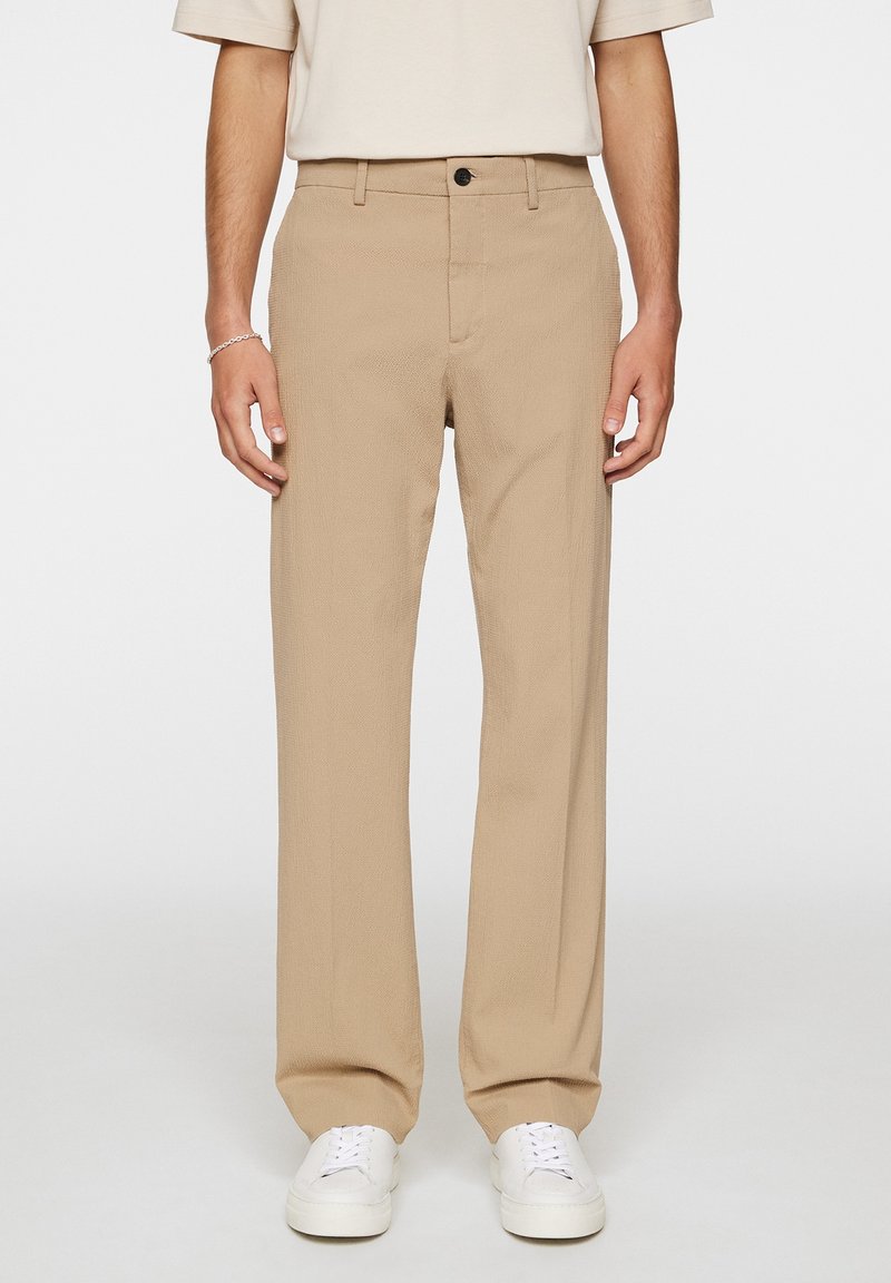 Beige Hosen aus strukturiertem Stoff, mit geradem Bein, frontalen Taschen und einem einzelnen Knopfverschluss. Getragen mit weißen Turnschuhen.