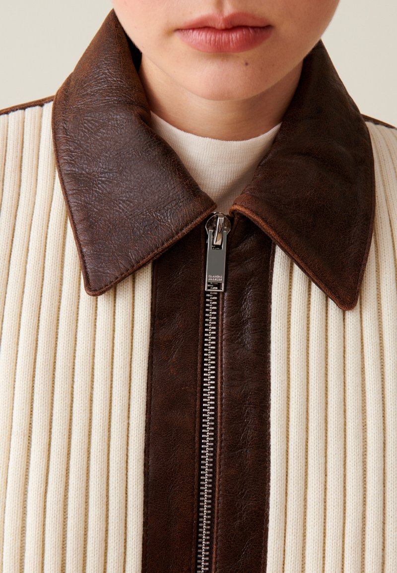 Brauner Lederkragen und Reißverschluss akzentuieren eine cremefarbene, gerippte Stoffjacke mit strukturiertem Design und kontrastierenden Paneelen.