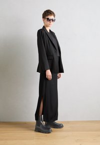 Černý přizpůsobený blazer přes černé šaty s bočními rozparky, doplněné černými Chelsea botami. Model má na sobě velké sluneční brýle. Neutrální pozadí.